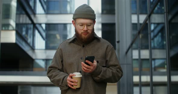 A Bearded Man is Typing on a Mobile Phone While Standing Near Office Building