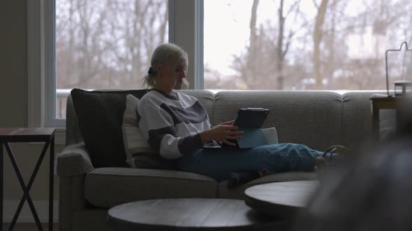 Woman sitting on the couch in front of a bay window, drinking coffee while scrolling on a tablet wit