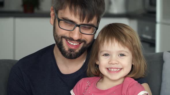 Father with Daughter Sit on Sofa at Home, Look at Camera, Smile and Laugh. Happy Family Concept.