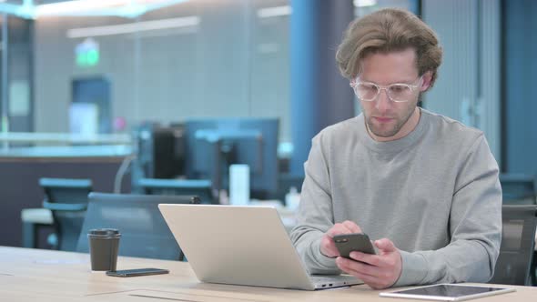 Young Businessman with Laptop Using Smartphone at Work