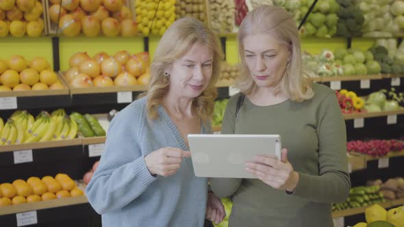 Portrait of Positive Caucasian Senior Women Using Tablet in Grocery. Two Housewives Standing Between