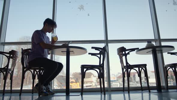 a Young Guy is Sitting in a Cafe By the Big Window Drinking Juice and Having Fun on the Internet