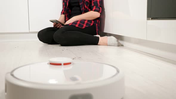 Unrecognizable Woman with Phone Sits on the Floor While the Robot Vacuum Cleaner Cleans the Floor