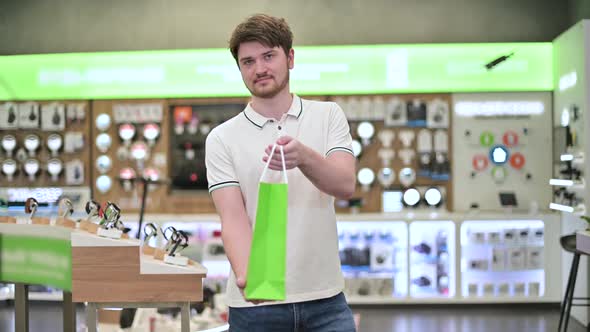 Young salesman in electronics store puts goods into paper bag and holds out