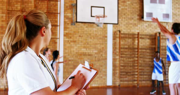 Female coach writing on clipboard while students playing in basketball court