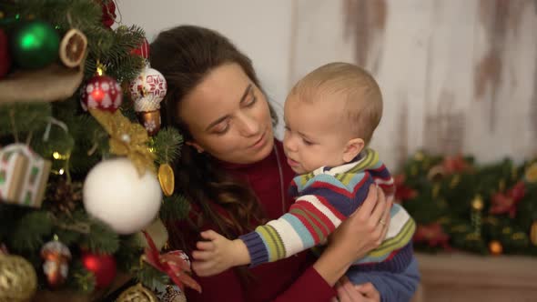 Pretty Young Woman Shakes the Baby in Her Arms Standing Near Christmas Tree and Showing Bright Toys