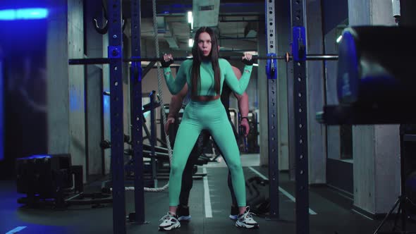 Woman in Sports Costume Trains Her Butt with Dumbbell on Her Shoulders Under the Supervision of a