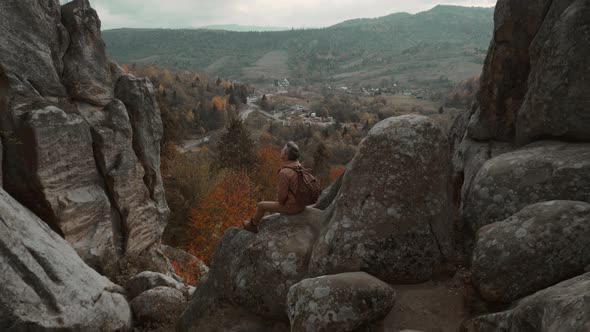 Rear View Man Hiker in Casual Brown Wear with Backpack Sits on Edge of Cliff Enjoys a Beautiful