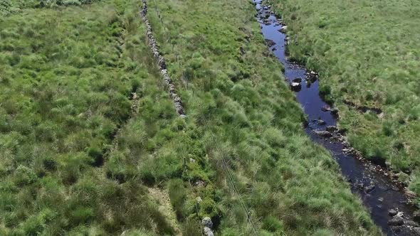 Mid shot aerial tracking forward over an old stone wall and along a river in the grassy moorland of