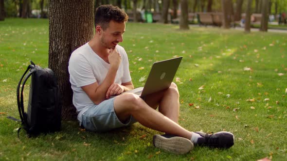 A Young Man on the Grass in the Park Sits Under a Tree and Has Fun Chatting on the Internet