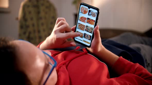 Young Man Lying on the Bed and Scrolling Pictures of Pizza on His Phone