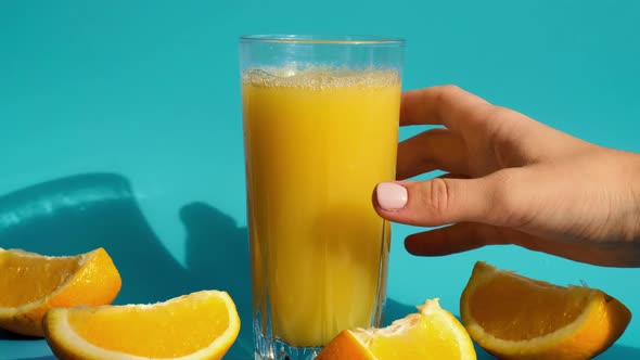 Female Hand Put and Take Glass of Fresh Squeezing Orange Juice with Sliced Fruits on Blue Background