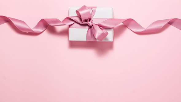 White gift box with a purple ribbon bow on pink background at the top. Flat lay top view stop motion