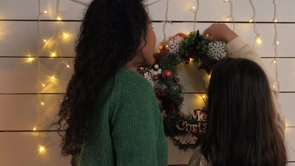 Mom and Daughter Hang a Christmas Wreath on the Wall Back View