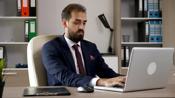 Attractive Young Businessman in the Office Types on the Computer Then Looks in a Folder with Charts