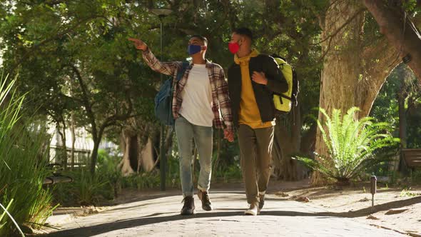 Two happy mixed race gay male couple walking and talking in park with backpacks