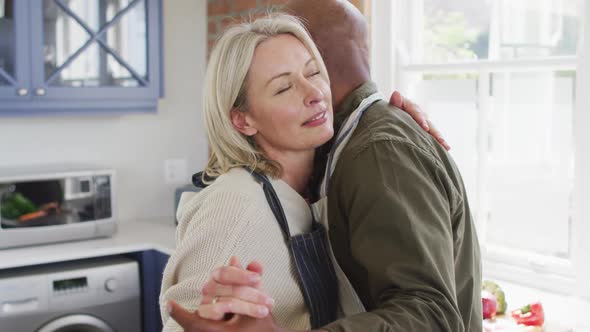 Mixed race senior couple wearing aprons dancing together in the kitchen at home