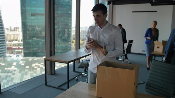 Young Employee Unpack Box with Docs and Items. His Colleagues Walk Near in Modern Office