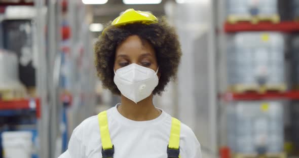 Female African Worker in Safety Helmet Wearing Face Protective Mask Walking in Warehouse