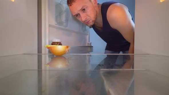 man opens the refrigerator and looks inside, there is an onion on the shelf.