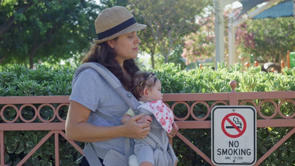 Woman and Baby in Carrier Standing Next to No Smoking Sign at Park