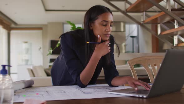 Mixed race woman by desk at home using laptop looking through blueprints