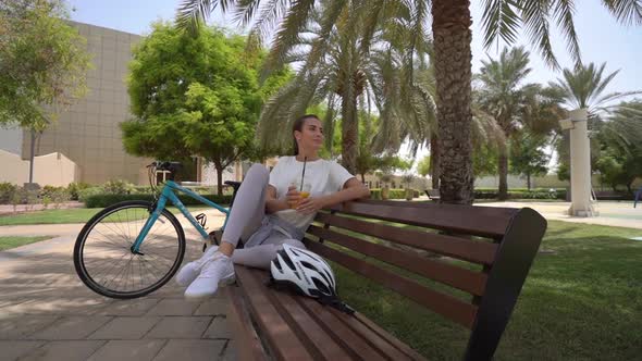 Young Woman Sits on Benches in the Park and Drinks Juice