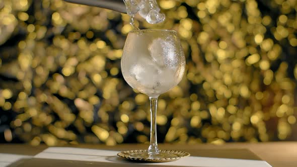 Ice Cubes are Being Dropped to the Cocktail Glass in Slow Motion on the Golden Sparkle Background