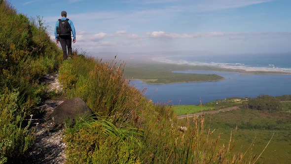 Man with drone backpack hiking a track on the side of a steep mountain-side with breathtaking views