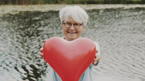 Senior Woman Holding Heart Shaped Balloon Near the River. Love and Care Concept