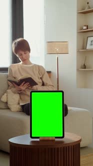 A Woman Reading a Book Sitting on a Sofa in Front of a Green Screen Tablet