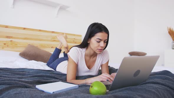 Сheerful Young Woman Working at Home Using Laptop While Laying in Bed