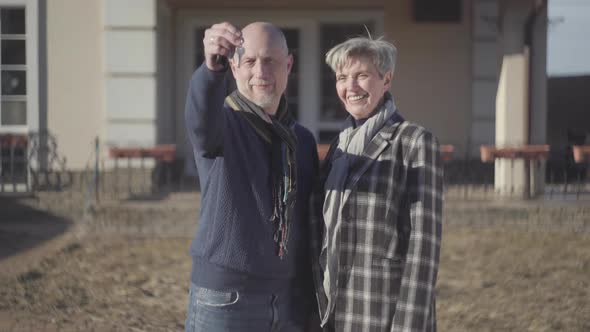 Happy Senior Couple, Bald Man and Woman with Grey Short Hair in Coat and Scarf Stand Near House