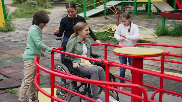 Cheerful Children Spinning Disabled Girl in Wheelchair on Merry Go Round Outdoors
