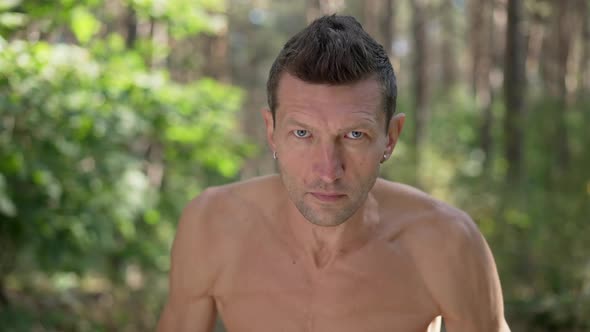 Zoom in to Serious Topless Sportsman Looking at Camera Standing in Sunshine in Forest