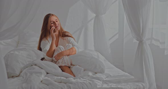 Cheerful Girl Talking on the Phone with Her Friend in the Bedroom, Joyful Morning. Happy Woman Hangs