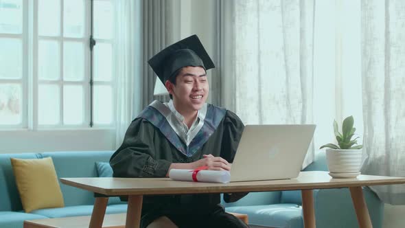 Happy Asian Man Wearing A Graduation Gown And Cap Use Laptop Computer In Living Room