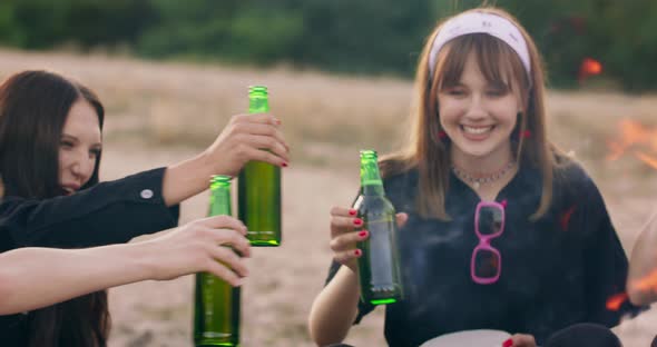 A Group of Young Adult Friends Toasts with Beer Bottles Outside Sitting Around a Campfire