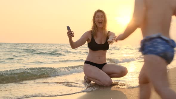 Beautiful Slender Blonde Mother in Black Swimsuit is Calling Her Baby on Beach at Sea or Ocean at