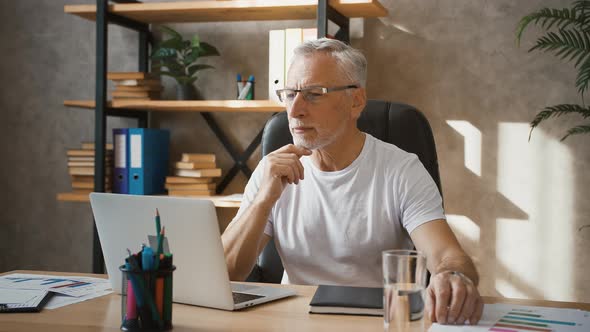 Grayhaired Businessman is Looking Thoughtful Sitting at Desk and Typing on His Laptop While Working