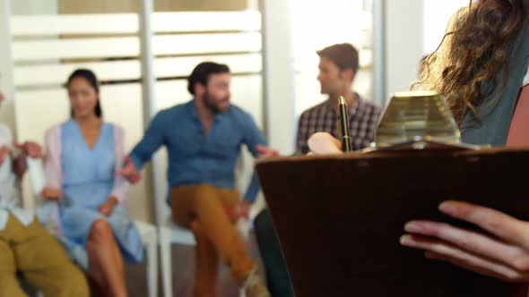 Businesswoman writing on clipboard while colleagues discussing in background