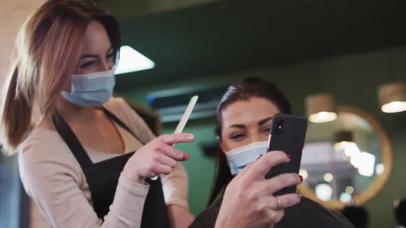 Female hairdresser and female customer wearing face masks looking at smartphone at hair salon