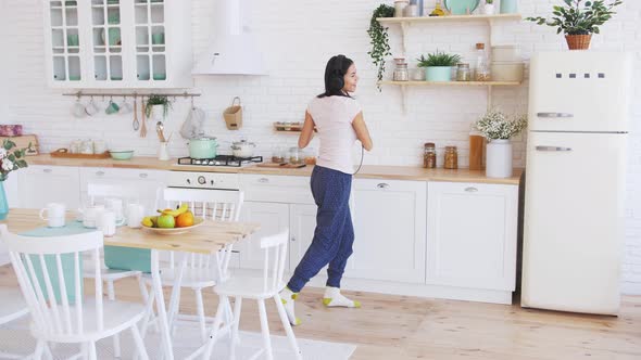 Young Happy Beautiful Woman Dancing in Kitchen Wearing Pajamas and Headphones and Listening to Music