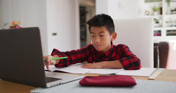 Asian boy at home, sitting at table using laptop for online school lesson