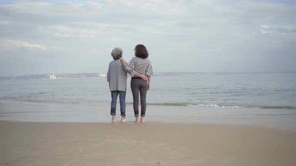 Two Smiling Women Hugging on Sandy Beach and Pointing at Distance