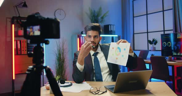 Businessman in Suit Sitting in front of Camera on Tripod in Evening Office and Explaining Chart