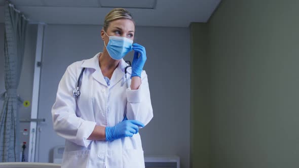 Thoughtful caucasian female doctor face mask and surgical gloves standing at hospital