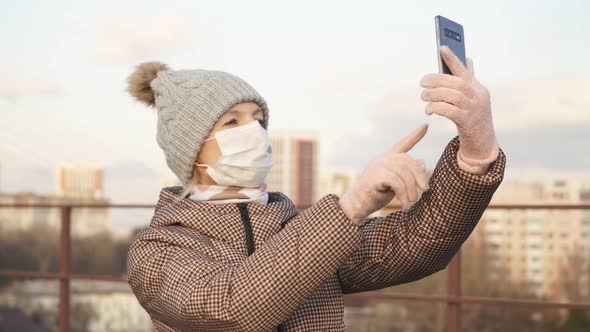 Young Woman in a Medical Mask on the Street Takes a Selfie on Her Smartphone Against the Background