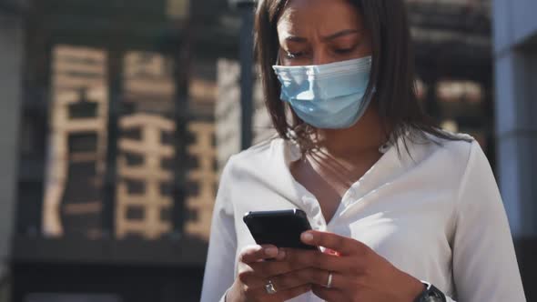 African american woman wearing face mask using smartphone in street