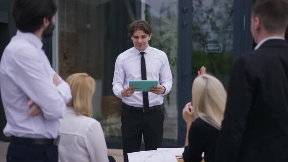 Confident Smiling Brunette Caucasian Man with Tablet Answering Colleagues Questions on Meeting